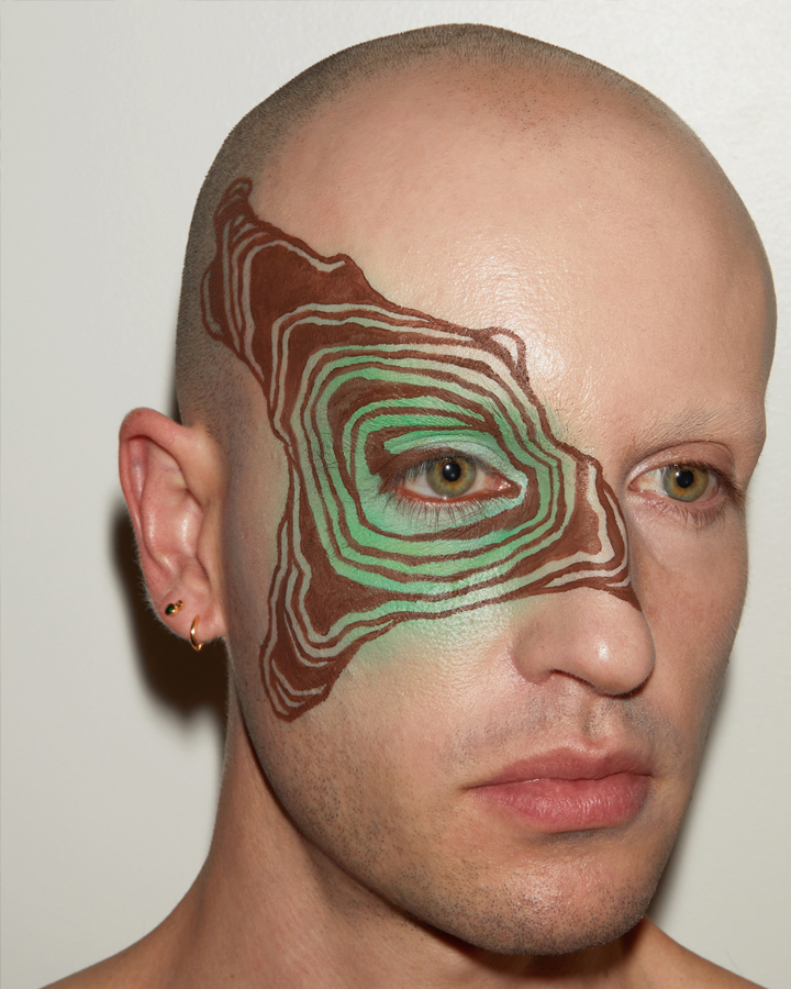 Jesse with graphic brown liner make up in concentric shapes extending out from one eye against a grey background